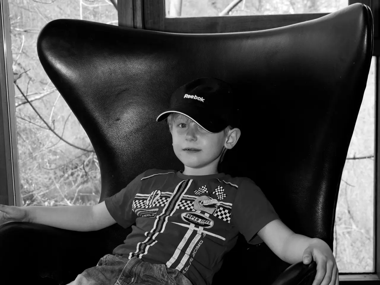 This image consist of a boy sitting in a black chair. He is wearing a black cap and blue jeans. In...