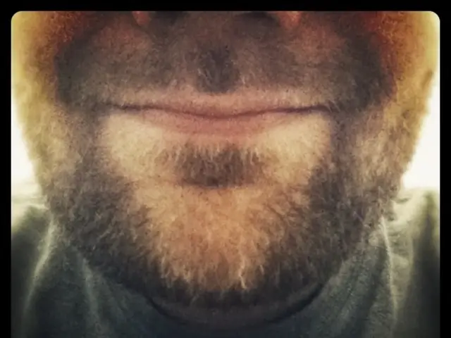 This image consists of a face of a man. He has a beard. He is wearing a black shirt.