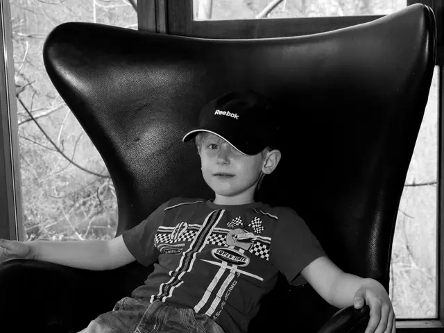 This image consist of a boy sitting in a black chair. He is wearing a black cap and blue jeans. In...
